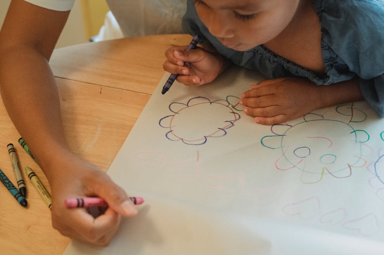 From above of crop anonymous female drawing with little daughter on paper with color wax crayons
