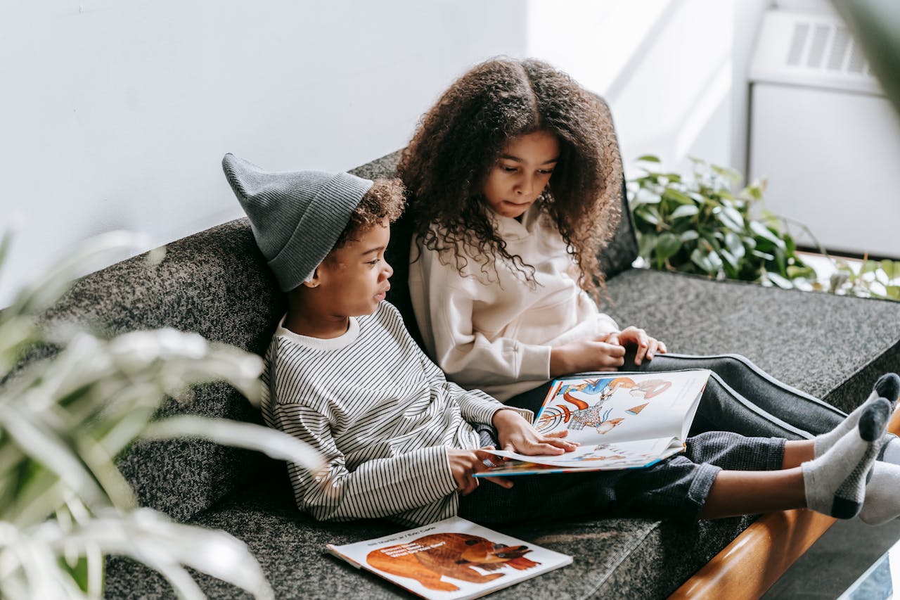 Two children enjoy reading storybooks together on a comfortable sofa in a bright indoor setting.