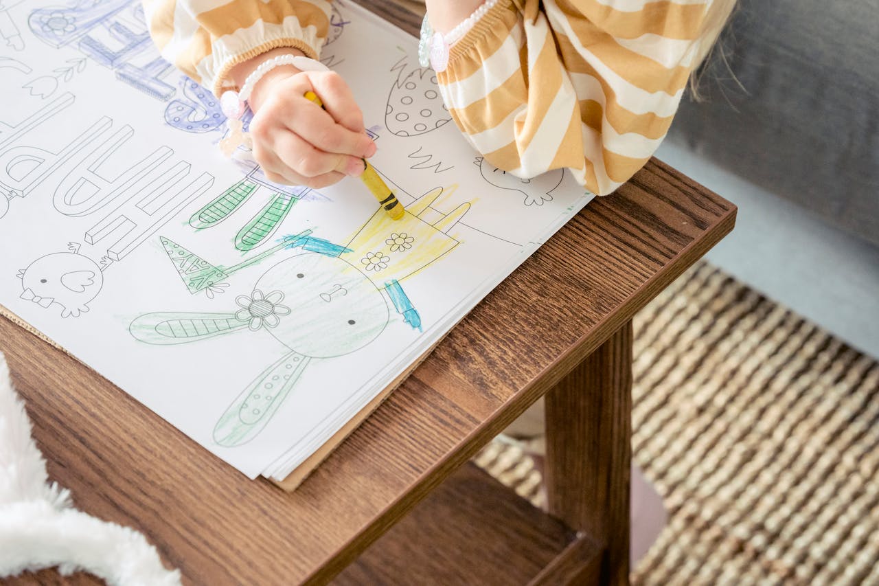 From above of crop anonymous little girl coloring bunny with colorful wax crayon on greeting poster for Easter