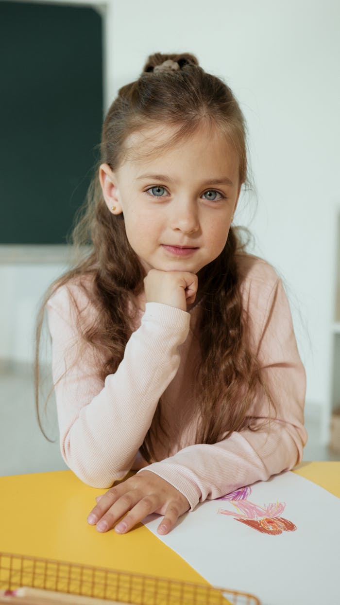 Adorable girl with long hair and blue eyes, seated in a classroom, drawing on paper.
