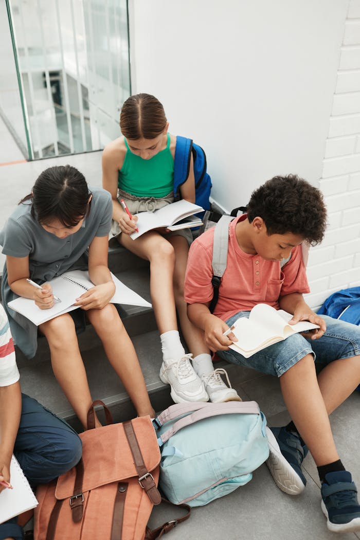 A group of students sitting on stairs, reading and writing with backpacks nearby.