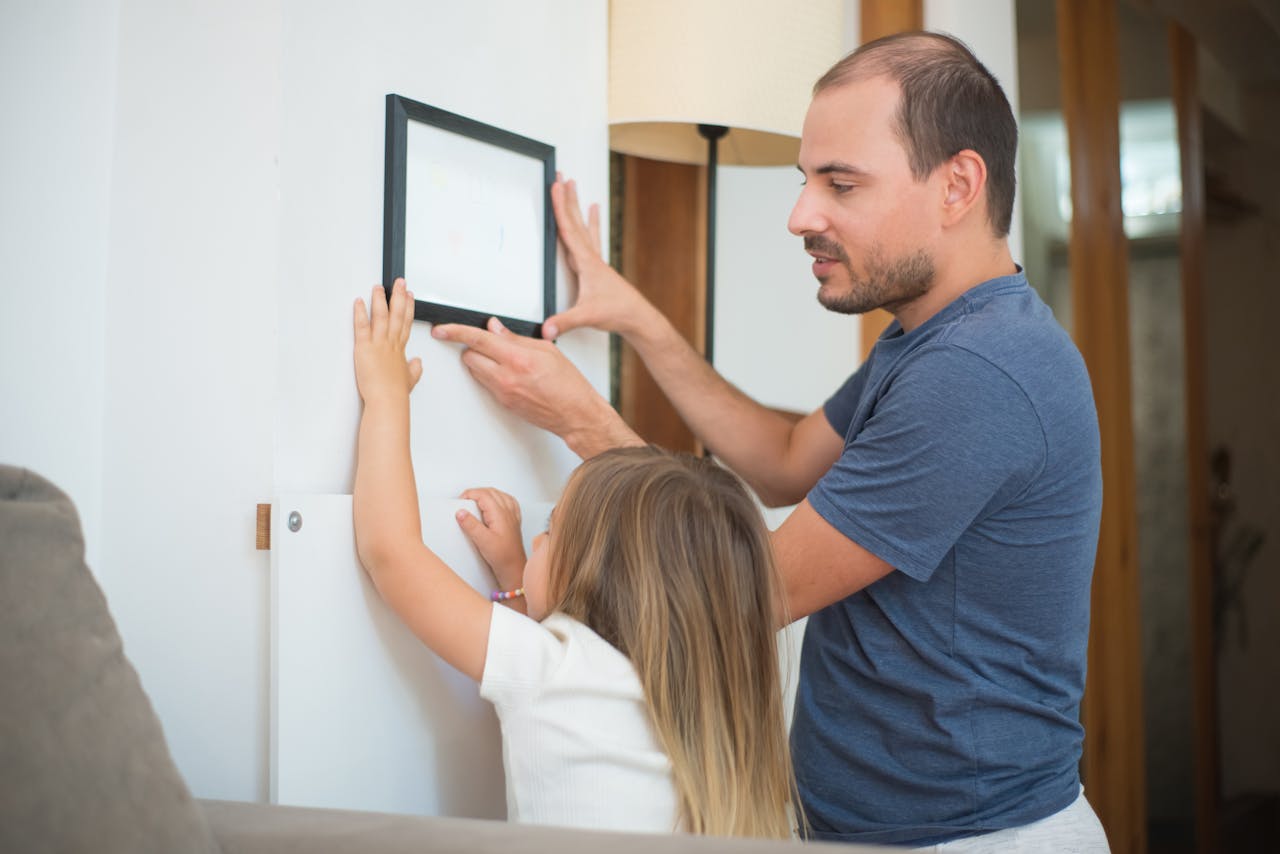 A father helps his daughter hang a picture frame on the wall, fostering teamwork and family bonding.